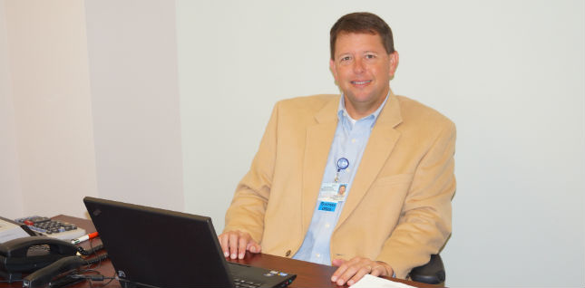 Derek Jackson - Business Office Manager of D.W. McMillan Memorial Hospital sitting at his desk smiling with his laptop open.