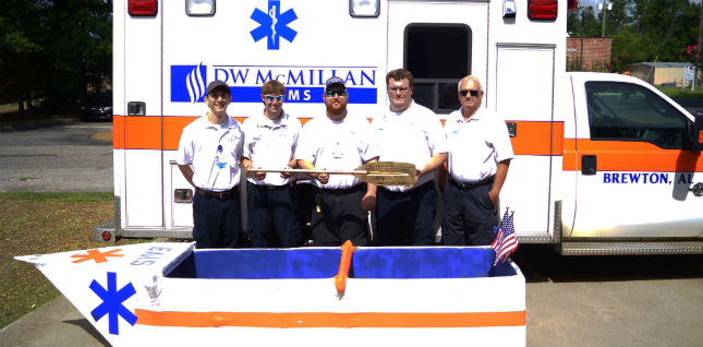 D.W. McMillan EMS staff standing on the side of an ambulance holding a paddle with a hand built little boat infant of them